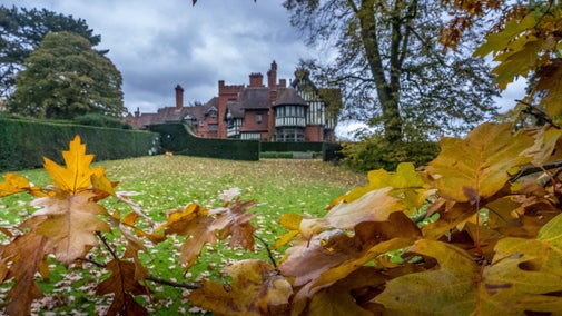 A view looking from the bottom of the paddock up towards Wightwick Manor in the distance. In the foreground are leaves which have changed colour with the season and are brown, orange and yellow.